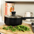 Black non-stick saucepan with glass lid on wooden board, with fresh herbs and green beans.