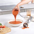 Hand blender mixing and pouring tomato soup into bowl, with fresh ingredients on cutting board