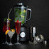 Black blender with mint, lime and ice, shown with three cocktails and bar tools on black background