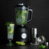 Black blender with jug of ice, lime and mint, cocktail glass and bar tools on black background