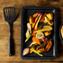 Roasting tray with assorted cooked vegetables beside black handled spatula on wooden surface