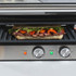 Stainless steel panini press with open grill plates, front-facing controls visible.