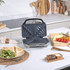 Electric sandwich maker open on kitchen counter, next to plate with toasted sandwich and salad.