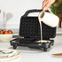 Black waffle maker on kitchen counter with batter being poured in