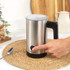 Hand pouring frothed milk from stainless steel jug into iced coffee, hot latte in background