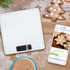 White digital kitchen scale on wooden surface with Christmas biscuits, smartphone, and hot chocolate.