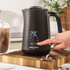 Black electric hot chocolate maker pouring drink, two topped mugs beside, kitchen setting