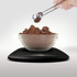 Glass bowl of cocoa powder on black digital kitchen scale, with scoop measuring powder above.