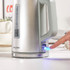 Silver electric kettle with visible water gauge and blue power switch being pressed