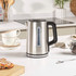 Stainless steel electric kettle with black handle, being switched on by a person's finger