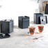 Set of three matte black square storage tins with lids, shown on a kitchen worktop with tea bags and tea glasses.