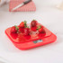 Red digital kitchen scale with five strawberries on top, shown on a white surface.