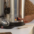 Person pressing on button of large silver hot water urn with teacup and teabag beside