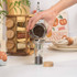 Person refilling glass pepper mill with black peppercorns beside wooden spice rack on kitchen counter