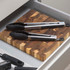 Pair of stainless steel kitchen tongs with black silicone tips on wooden board in drawer