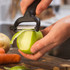 Black-handled peeler peeling a green apple, with potato and carrots on a wooden board.