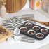 Six chocolate chip muffin batter portions in white paper cases in a non-stick tray on kitchen worktop.