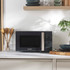 Grey microwave with silver handle on kitchen worktop, shelves and utensils around