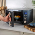Grey microwave on countertop with door open, bowl of food inside, hand holding handle
