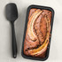 Non-stick black loaf tin with freshly baked banana bread and a black spatula beside it