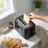 Black 2-slice toaster on kitchen counter, person inserting bread, toast and jam visible