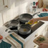 Set of three grey non-stick frying pans on hob, oil being poured into one pan.