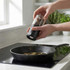 Hand using black Salter grinder to season food in frying pan on induction hob