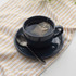 Black cup and saucer with tea bag and spoon on patterned cloth.