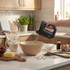 Handheld black electric mixer whisking batter in bowl, kitchen counter with baking ingredients.