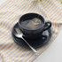 Black ceramic cup and saucer with tea bag and spoon on patterned cloth.