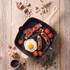 Square cream grill pan with cooked bacon, sausages, egg and tomatoes on wooden surface