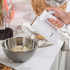 White electric hand mixer blending dough in metal bowl, kitchen worktop.