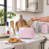Pink two-slice toaster on kitchen counter, hand adding seeded bread, matching kettle in background