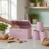 Pink metal bread bin with lid open, hand placing sliced bread inside, on kitchen counter