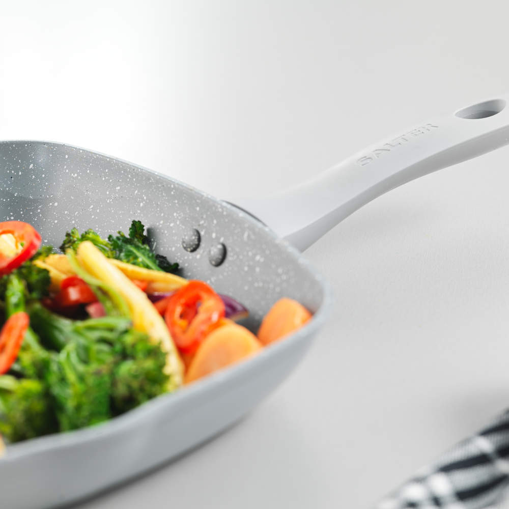 Silver frying pan with speckled coating, cooking mixed vegetables, angled on white surface