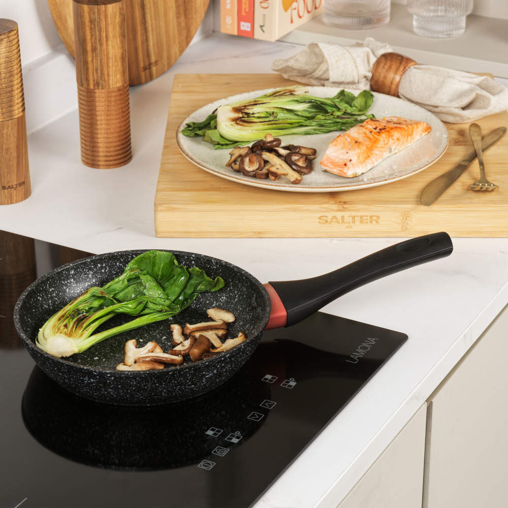 Non-stick frying pan with black handle on induction hob, cooking pak choi and mushrooms.