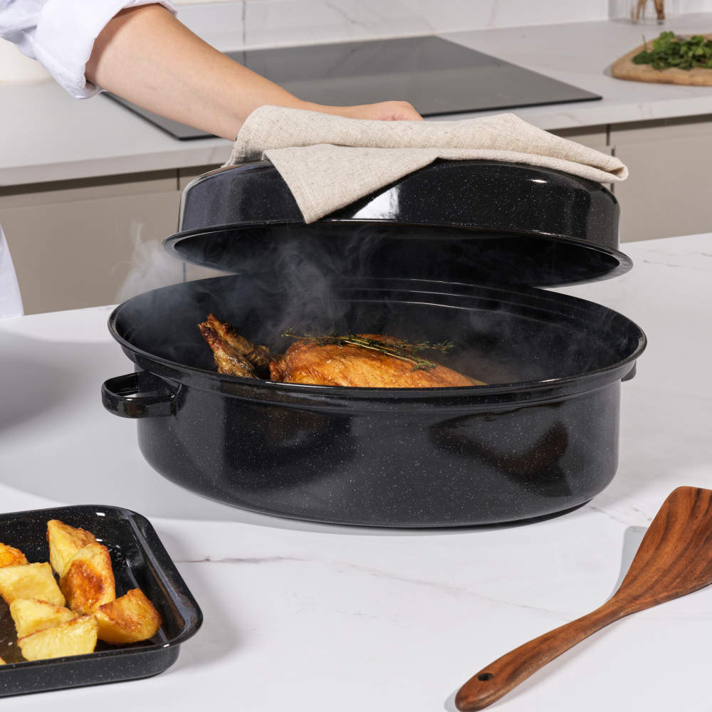 Black enamel roasting pan with lid, shown with cooked chicken and herbs inside on kitchen worktop.