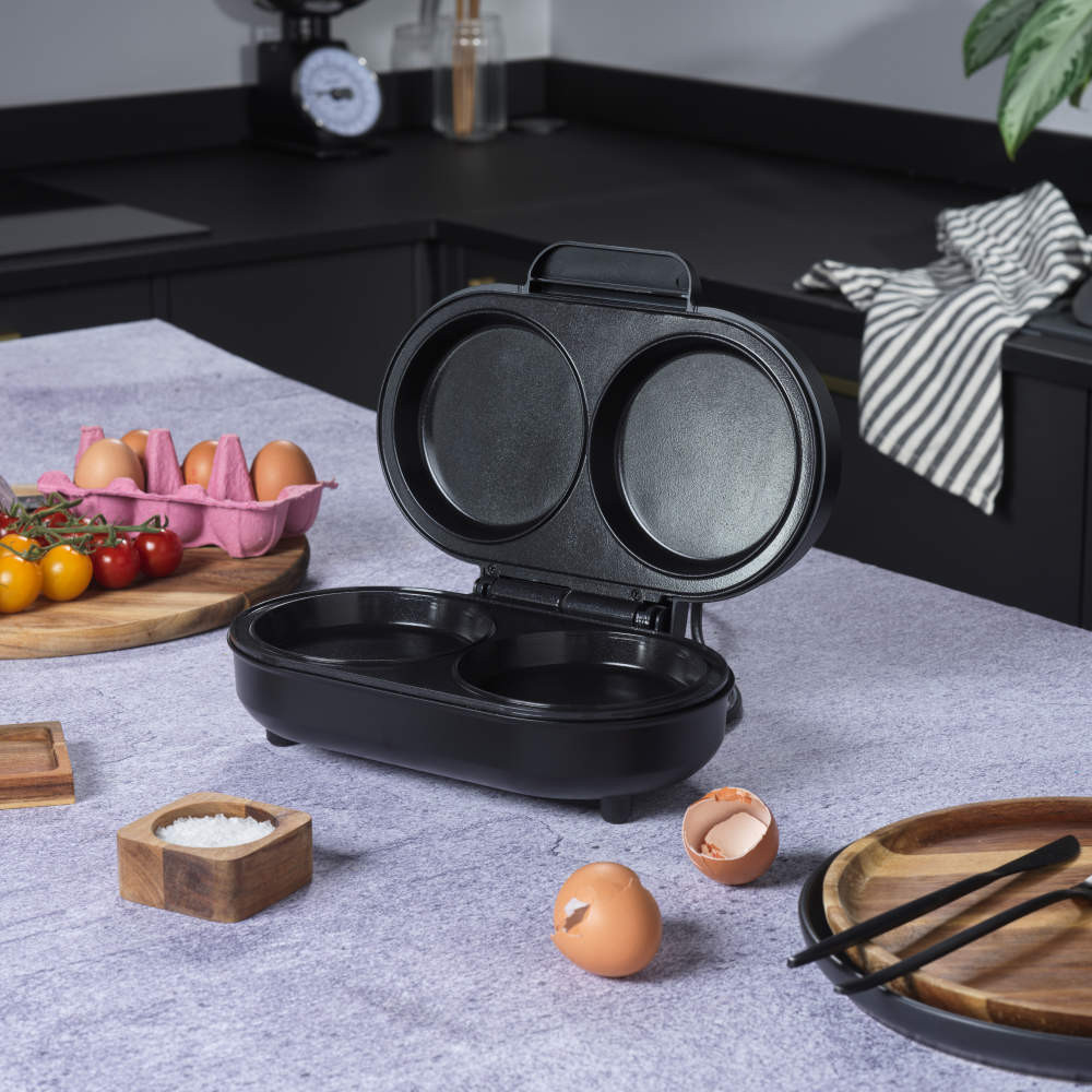 Black electric egg cooker with two round non-stick compartments, open and empty, front view