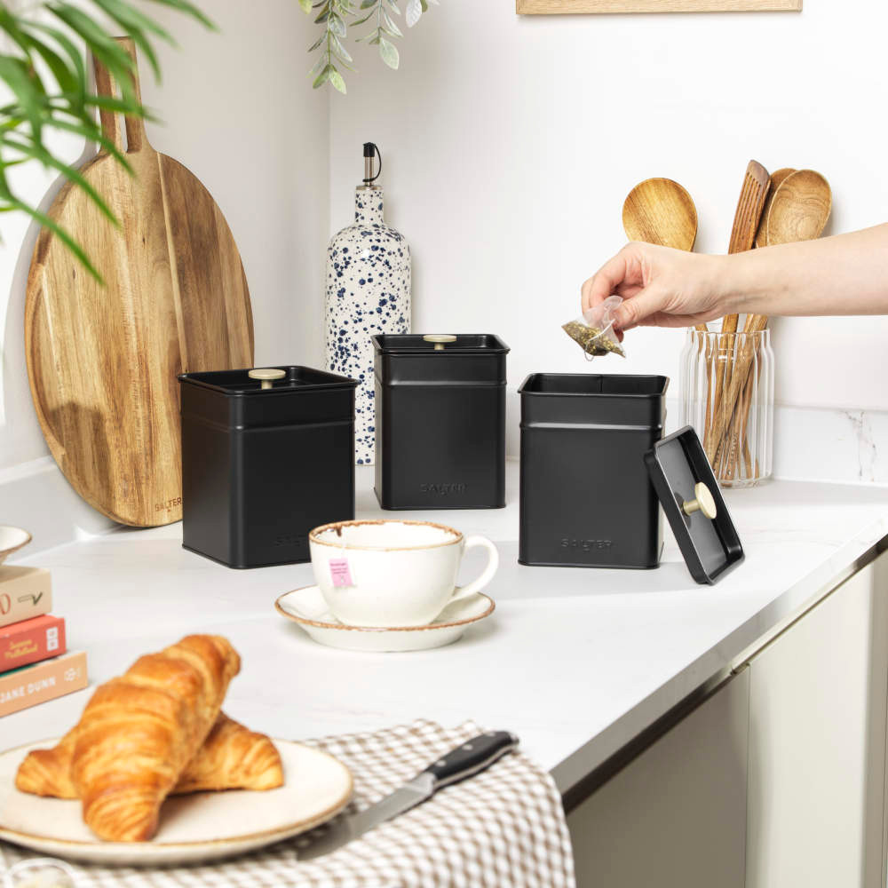 Set of black kitchen storage tins on worktop, one opened with hand holding tea bag