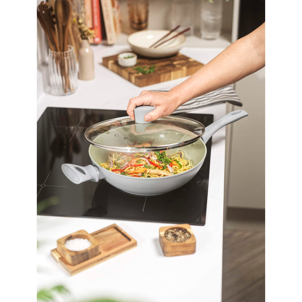 Light green non-stick wok with two grey handles and glass lid, shown at angle