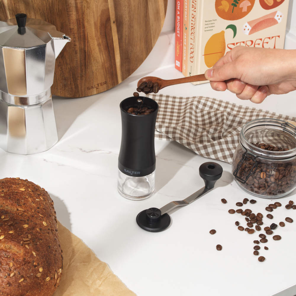 Black manual coffee grinder with side handle, clear base, shown on white background