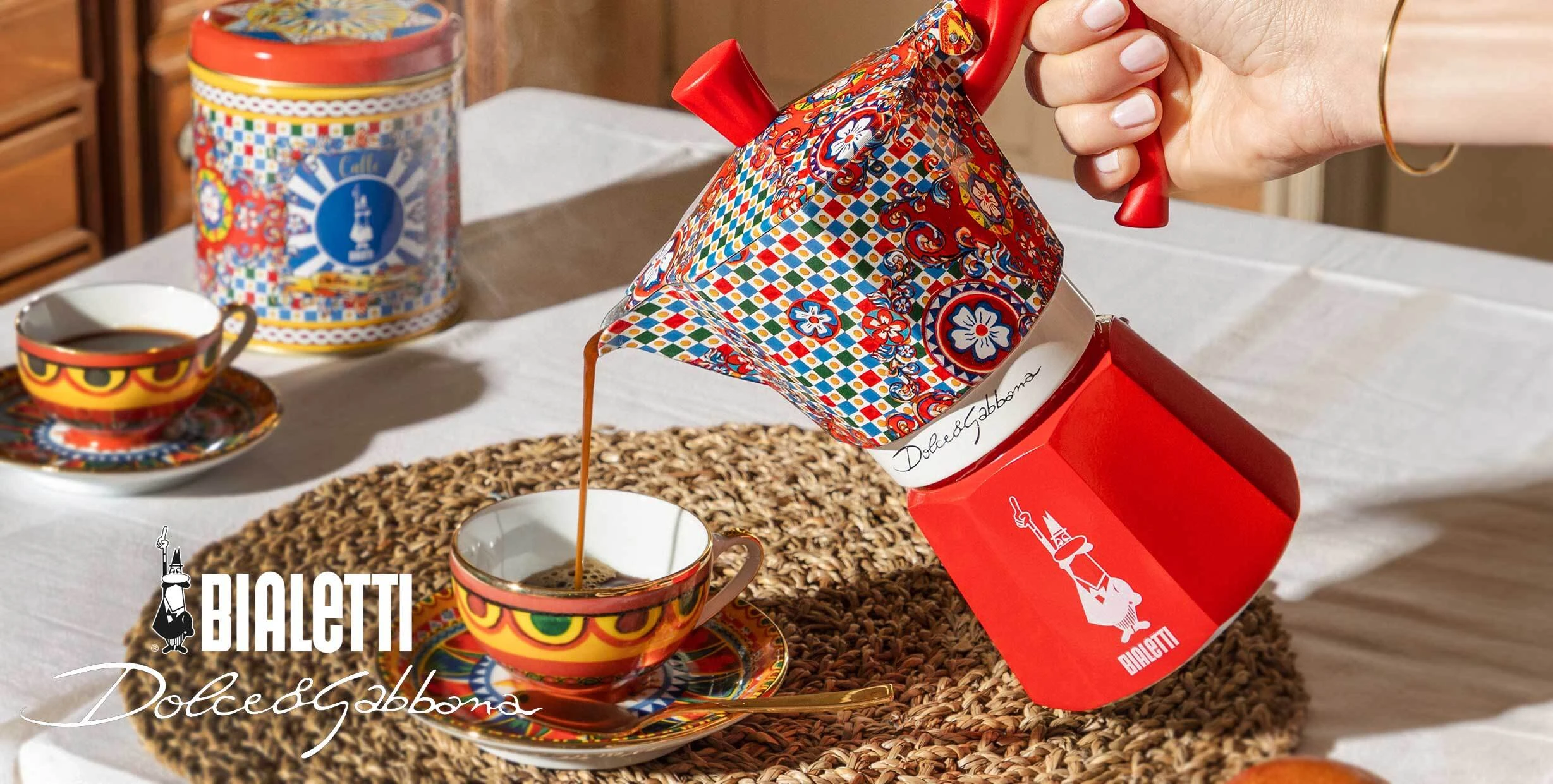 Bialetti Dolce & Gabbana moka pot pouring espresso into decorative cup, colourful Sicilian design stovetop coffee maker on table with matching espresso set