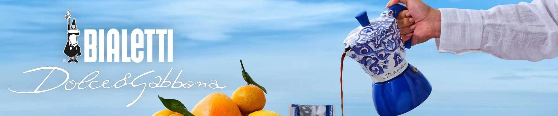 Bialetti Dolce & Gabbana blue moka pot pouring coffee, Italian stovetop espresso maker with ornate pattern, citrus fruits and coastal background