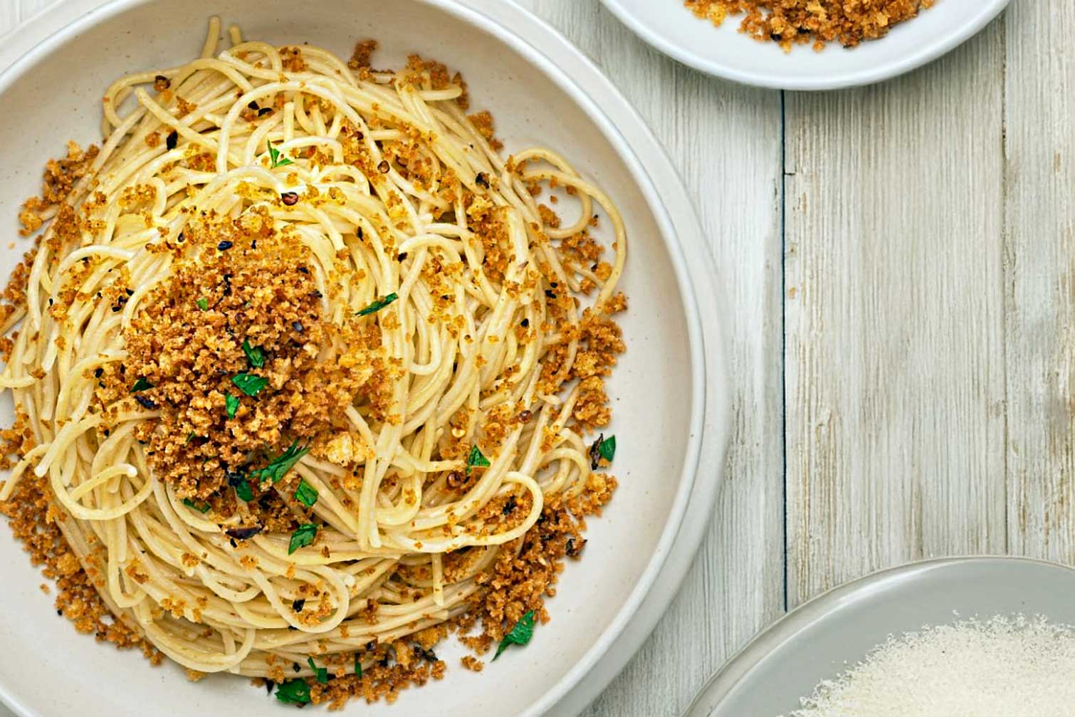 A plate of Lemon and Chilli Spaghetti made with Pastificio Liguori Spaghetti Quadrati, tossed in Hillfarm Cold Pressed Rapeseed Oil, topped with Maldon Garlic Salt pangrattato and fresh parsley – a vibrant summer pasta dish served on a rustic wooden table.