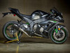 M4 Performance Exhausts M4 Tech1 Carbon Slip On Exhaust Kawasaki ZX-10R (11-20) with Titanium Cat Eliminator, mounted on a black Kawasaki ZX-10R motorcycle, in a showroom or garage setting.