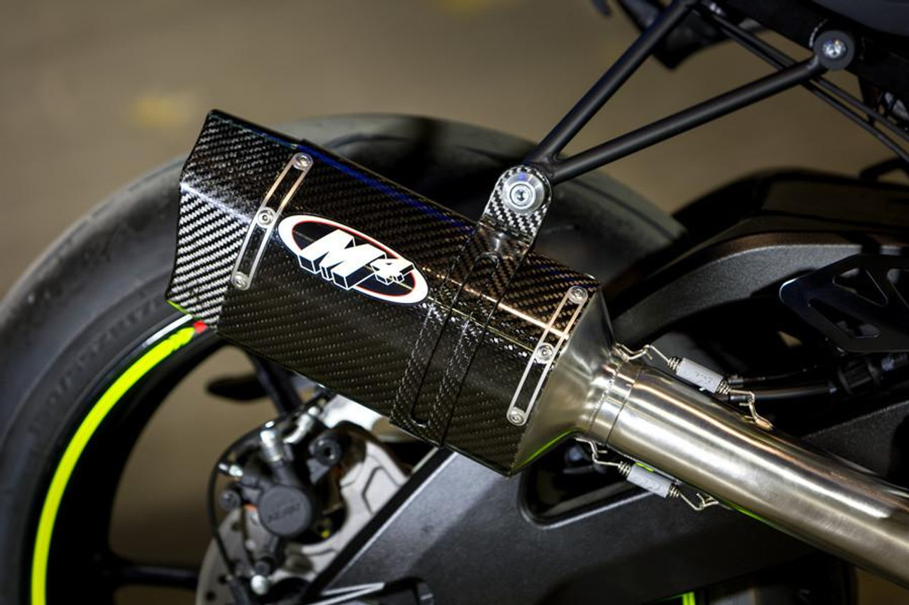 M4 Performance Exhausts M4 Tech1 Carbon Exhaust Suzuki GSXR1000 (17-25) w/Titanium Midpipe, close-up view showing the carbon fiber finish and logo on the exhaust with the motorcycle's rear wheel and suspension in the background.