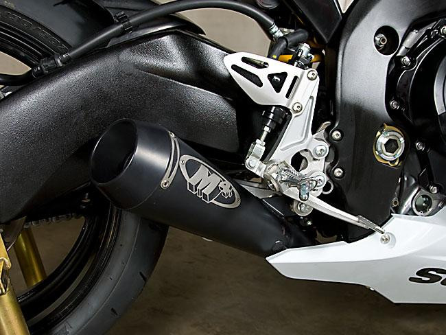 M4 Performance Exhausts M4 GP Black Slip On Exhaust Suzuki GSXR1000 (09-11), close-up view of the black exhaust pipe with skull logo, mounted on the motorcycle's black frame and rear wheel.