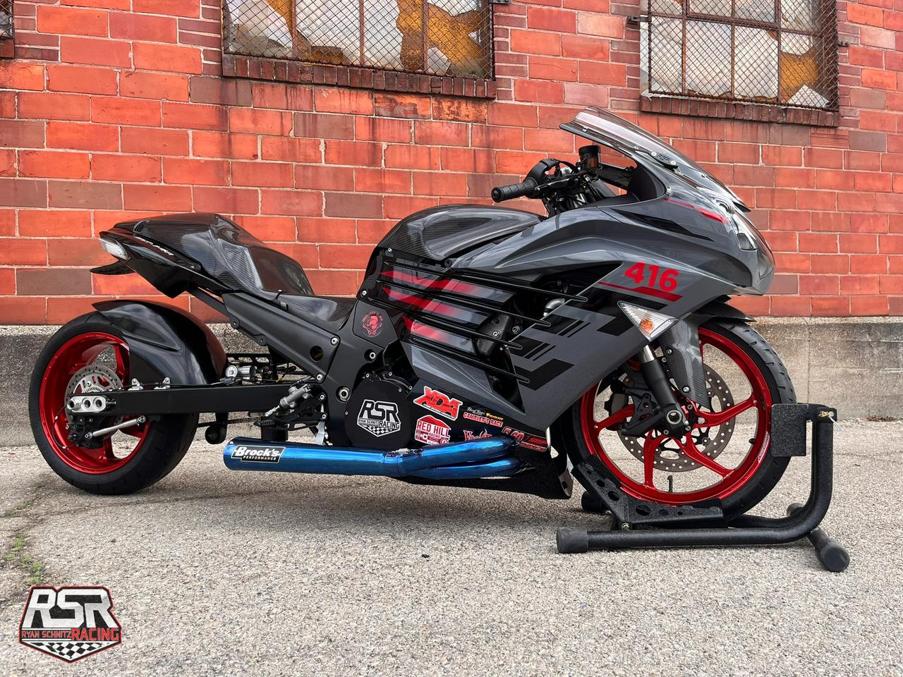 Montgomery Motorsports Carbon Fiber Low Profile Fuel Tank Kawasaki ZX-14R (12-26), black and gray motorcycle with red accents parked on a concrete surface against a brick wall background.