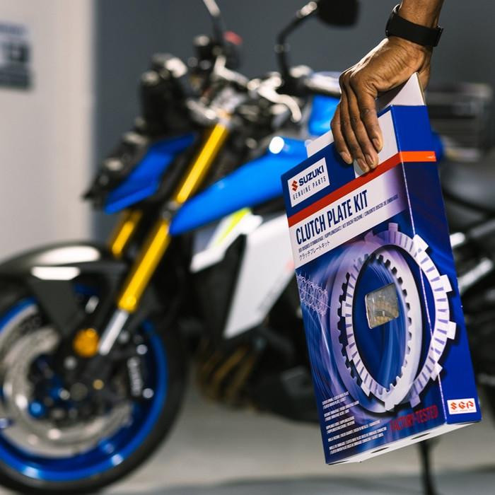 Suzuki OEM Complete Clutch Kit Suzuki GSXR600 (11-26), new clutch plate kit in packaging held by a person with a motorcycle in the background.