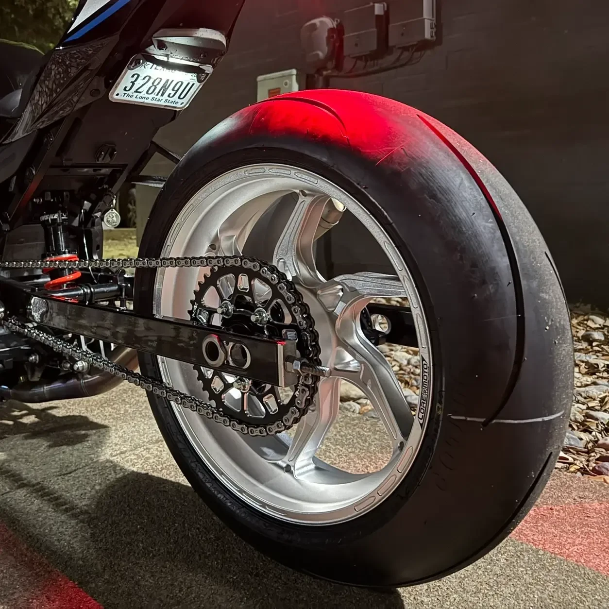 Vortex Motorcycle Parts Vortex V4 Lightweight Rear Sprocket, close-up view of the motorcycle's rear wheel and sprocket with chain, mounted on a sleek black motorcycle with a reflective tire, outdoor nighttime setting.